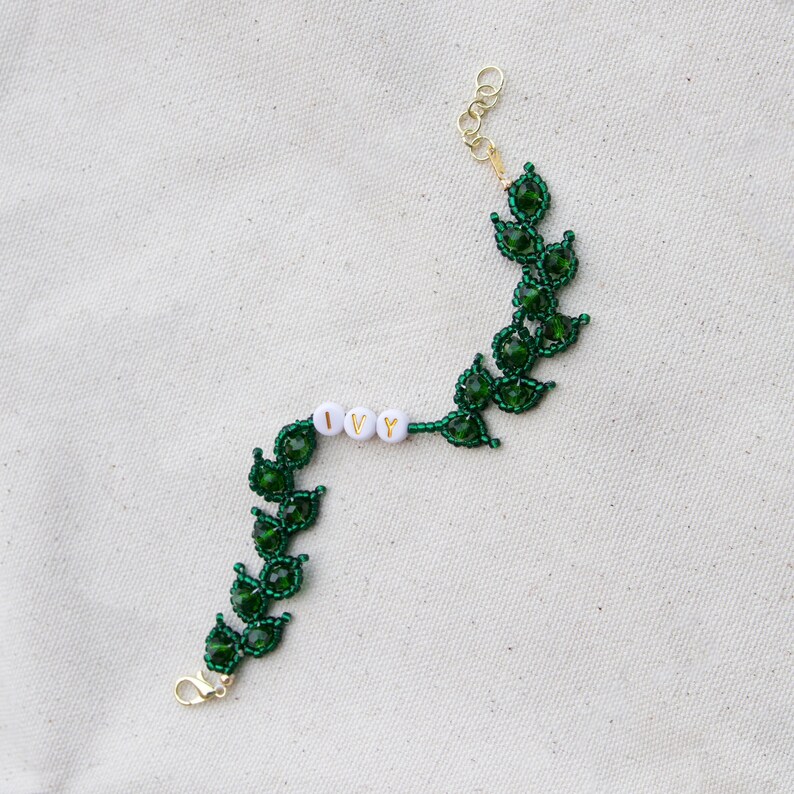 May include: A green beaded bracelet with a leaf design. The word "IVY" is spelled out in white letter beads. The bracelet has a gold-colored clasp and extender chain.