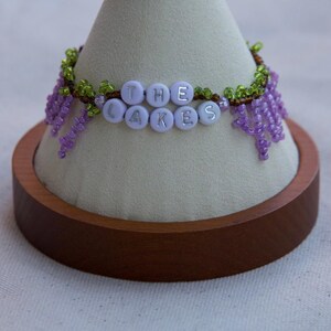 May include: A beaded bracelet with the words "THE LAKES" spelled out in silver letters on light purple beads. The bracelet features green and brown beaded accents and dangling purple beaded strands, displayed on a white cone-shaped stand.