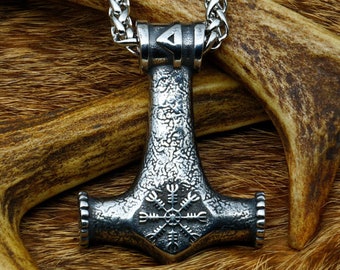 May include: A silver Mjolnir pendant with a Vegvisir symbol on the front, hanging from a silver chain. The pendant is resting on a piece of brown animal fur.