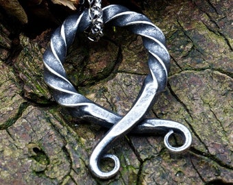 May include: A handcrafted, silver-colored metal pendant necklace. The pendant features a twisted, heart-shaped design with a looped bottom and a chain. The necklace is displayed on a textured, brown wooden surface.