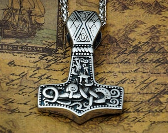 May include: A silver Mjolnir pendant with intricate details and a chain. The pendant is on a brown background with writing.