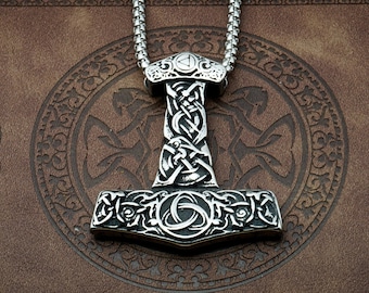 May include: A silver Mjolnir pendant necklace with intricate knotwork designs. The pendant is on a silver chain and is resting on a brown leather surface.