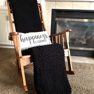 May include: A black chunky knit throw blanket draped over a wooden rocking chair with a white pillow that reads "happiness is homemade".
