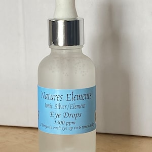 Puede incluir: Una botella de vidrio transparente con una tapa plateada y un gotero blanco. La botella está etiquetada con "Natures Elements Ionic Silver/Element Eye Drops 1300 ppm 2 Drops in each eye up to 6 times a day".