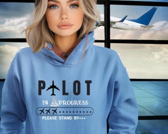 May include: A light blue hoodie with the text "Pilot in Progress" and a graphic of a plane taking off. The text "Please Stand By..." is below the graphic.