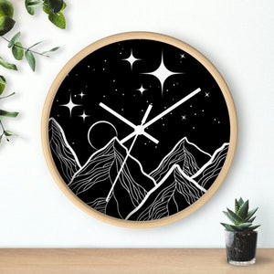 May include: A round wall clock with a light wood frame. The clock face is black with a white mountain range design, a moon, and stars. The clock hands are white.