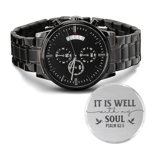 May include: A black wristwatch with a matching black band. The watch face has multiple sub-dials and a date display. A separate silver-colored disc with the text "IT IS WELL with my SOUL PSALM 62:5" is also visible.
