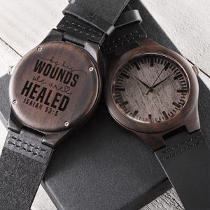 May include: Two wooden watches with black leather straps. One watch face displays the time, while the other features the engraved text "by his wounds we are healed" and the Bible verse Isaiah 53:5. The watches have a dark brown wood grain finish.