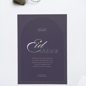 Könnte beinhalten: Eine lila Eid Mubarak-Grußkarte mit dekorativem Rand und Kalligraphie. Die Karte enthält den Text "Eid Mubarak" und eine Botschaft, die Freude, Frieden und Segen wünscht. Ein Zweig Blätter liegt über der Karte.