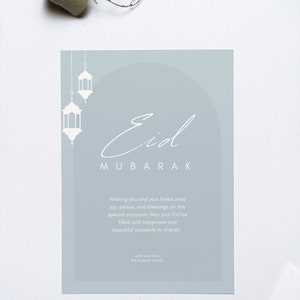 K&ouml;nnte beinhalten: Eine hellblaue Karte mit dem Text "Eid Mubarak" in wei&szlig;er Kalligraphie. Die Karte zeigt eine wei&szlig;e Silhouette eines Bogens mit drei h&auml;ngenden Laternen. Der Text "Wishing you and your loved ones joy, peace, and blessings on this special occasion. May your Eid be filled with happiness and beautiful moments to cherish. with love from the Hussain Family" ist in wei&szlig; gedruckt.