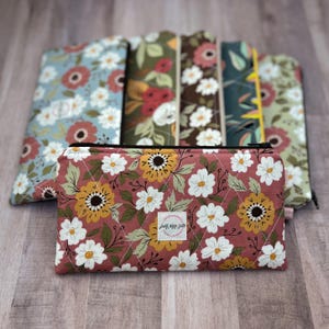 May include: A collection of floral print pouches in various colors, including a quilted, dusty rose pouch with white and yellow flowers. The pouches have a black zipper and a small label that reads "just sew cute". The background includes other floral print pouches.