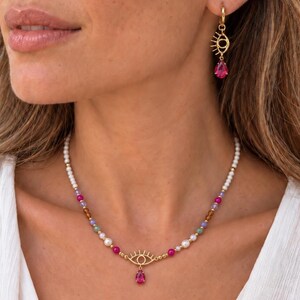 May include: A gold-toned necklace and matching earring set featuring an eye-shaped charm with a dangling teardrop-shaped fuchsia gemstone. The necklace is composed of white beads, small gold-toned spacers, and multicolored accent beads.