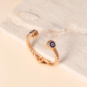 May include: A gold-toned metal hinged cuff bracelet featuring a twisted rope texture. The two ends of the bracelet are capped with circular ornaments, each displaying a blue, white, and black evil eye motif centered on the rounded surface.