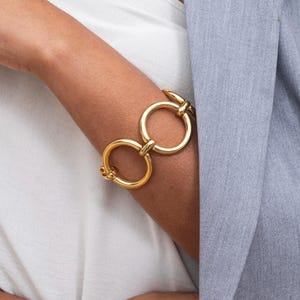May include: A gold-toned bracelet featuring two large, interlocking circular links connected by a smaller central bar. The bracelet has a polished, reflective surface and a toggle-style clasp mechanism visible on the side of the wrist.