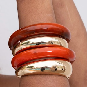 May include: A stack of four chunky bangles worn on the arm. The set consists of two smooth, polished gold-tone metal bangles alternating with two translucent, deep amber-colored resin bangles, all featuring a rounded, thick, and circular profile.