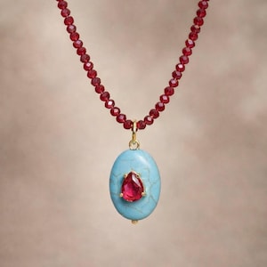 May include: A necklace featuring a strand of small, faceted red beads. The pendant is a light blue oval stone with a gold setting, holding a teardrop-shaped red gemstone. The necklace has a gold clasp.