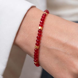 Op de afbeelding: Een rode kralenarmband met goudkleurige vierkante accenten. De armband is gemaakt van kleine, facetgeslepen rode kralen geregen met goudkleurige vierkante kralen. De armband wordt om een pols gedragen, met een wit overhemd op de achtergrond.