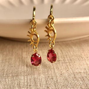 May include: A pair of gold-colored earrings with a sun and eye design. Each earring features a dangling, oval-shaped, red gemstone. The earrings are displayed on a neutral-colored surface.