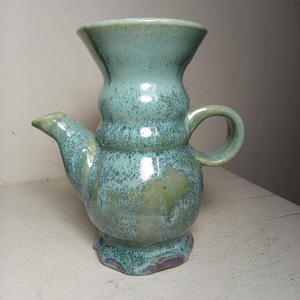 May include: A handcrafted ceramic teapot with a unique design. The teapot features a teal glaze with green and blue accents. It has a spout, handle, and a flared top. The base has a decorative, scalloped edge. A functional and decorative piece for the home.