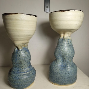 May include: Two tall ceramic goblets with a distinct two-tone design. The upper bowl section is a smooth, off-white cream color with a dark brown rim, while the lower base is a textured, speckled blue glaze with an irregular, organic shape.