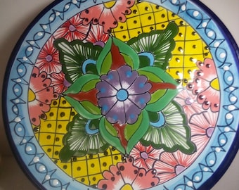 May include: A colourful ceramic plate with a blue rim and a floral design. The plate features a central flower with green leaves and a yellow background with a grid pattern.