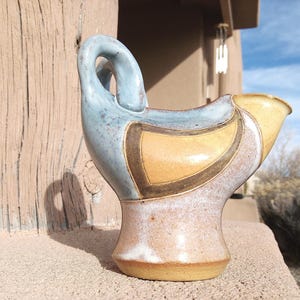May include: A handcrafted ceramic pitcher with a unique bird-like design. The pitcher features a light blue handle and accents, with a yellow and brown body. The base is a warm, earthy tone. The piece is displayed outdoors.