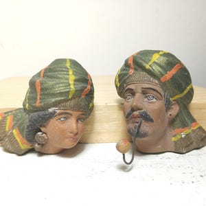 May include: Two vintage ceramic head sculptures. One depicts a woman, the other a man with a mustache and a pipe. Both wear turbans with green, yellow, and orange stripes. The sculptures are painted with earthy tones.