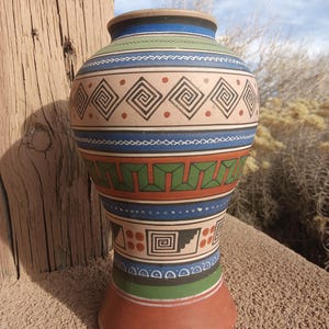 May include: A handcrafted, terracotta vase with a wide body and flared base. The vase features geometric patterns in shades of blue, green, red, and black against a tan background. The design includes repeating square and spiral motifs, creating a Southwestern aesthetic.