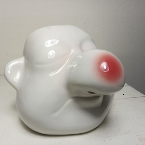 May include: A white ceramic pig-shaped container with a glossy finish. The pig has a prominent snout with a red-painted nose, small ears, and a rounded body. The container is likely a piggy bank or decorative item.