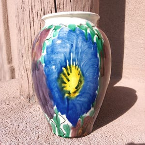 May include: A ceramic vase with a large blue flower design. The flower has a yellow center and is surrounded by green leaves and purple and red accents. The vase has a white rim and a brown base.
