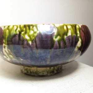 May include: A ceramic bowl with a pedestal base. The bowl features a glossy glaze with colors of green, purple, and blue. The bowl's rim has a green and white pattern, with purple and blue hues on the sides.