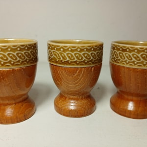 May include: Three egg cups featuring a polished wooden base with a visible grain pattern and a glazed upper rim in a tan color. The upper rim is decorated with a repeating white embossed rope-like pattern encircling the top of each cup.