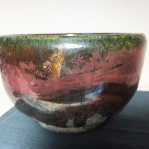 May include: A handmade ceramic bowl with a unique glaze. The bowl features a blend of colors, including green, red, and brown, creating a marbled effect. The rim has a dark green hue, while the body transitions to red and brown tones. The bowl's base is a lighter, unglazed color.