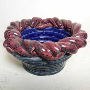 May include: A ceramic bowl with a unique design. The rim features a braided pattern in a deep burgundy color with teal speckles. The interior is a vibrant blue, and the base is a darker blue with horizontal ridges. A decorative piece.