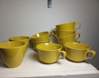 May include: A collection of mustard yellow teacups with handles. Some cups are stacked, others are arranged individually. The cups are made of a glossy material and sit on a white surface.