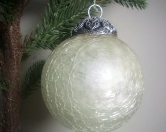 May include: A white, crackled glass Christmas ornament with a silver metal top. The ornament is hanging from a green artificial Christmas tree.