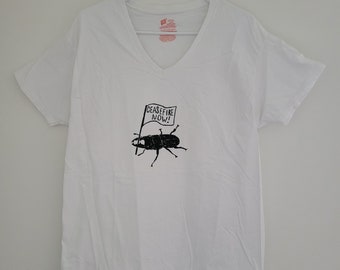 May include: White V-neck t-shirt with a black and white graphic of a beetle holding a flag that says "Ceasefire Now!"