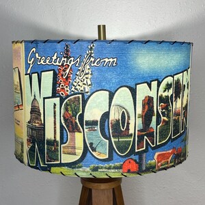 May include: A lampshade with a vintage postcard design featuring the text "Greetings from Wisconsin". The postcard design includes images of the Wisconsin State Capitol building, a lake, and a farm scene. The lampshade is made of fabric and has a black stitched trim.