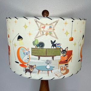 May include: A white lampshade with a black trim features a mid-century modern design with illustrations of dogs, furniture, and plants. The lampshade has a white background with gold stars and a black trim.