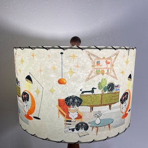 May include: A white lampshade with a black trim features a mid-century modern design with a repeating pattern of dogs reading books, a clock, and a potted plant. The lampshade is decorated with gold stars.