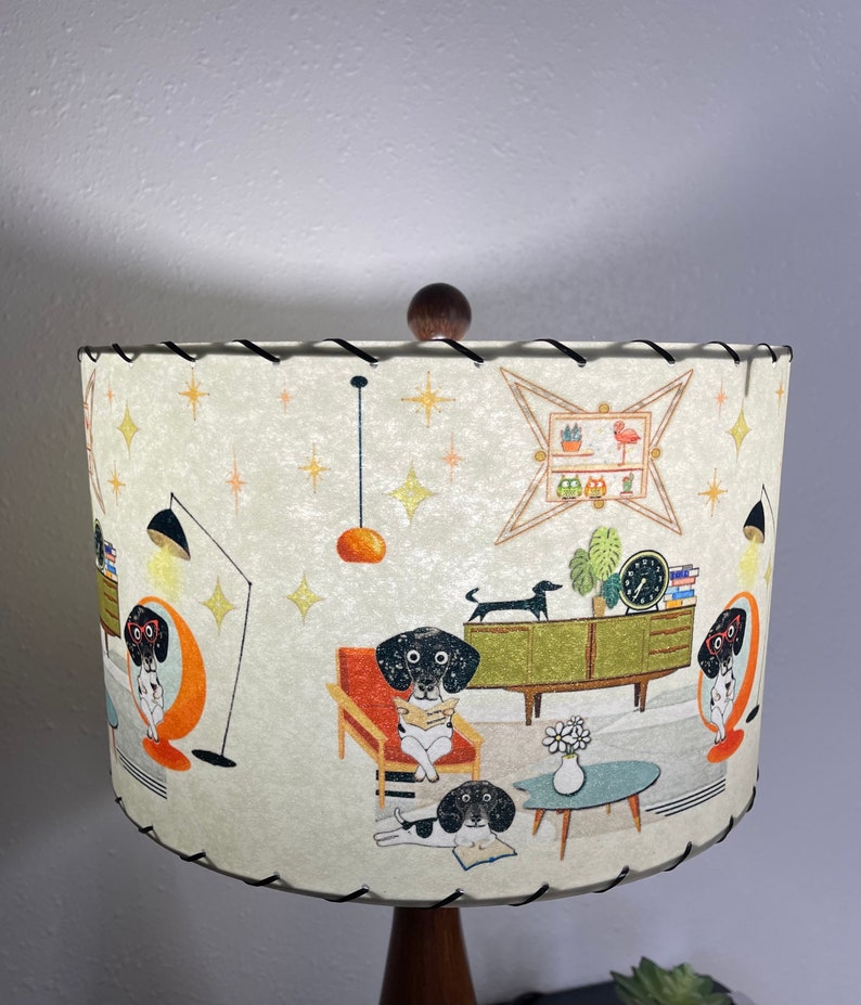 May include: A white lampshade with a black trim features a mid-century modern design with a repeating pattern of dogs reading books, a clock, and a potted plant. The lampshade is decorated with gold stars.