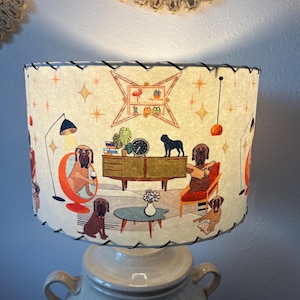 May include: A cream-colored lampshade with a retro dog-themed design. The shade features illustrations of dogs in a mid-century modern living room setting, with furniture, plants, and a clock. The shade is trimmed with black stitching and sits atop a ceramic base.