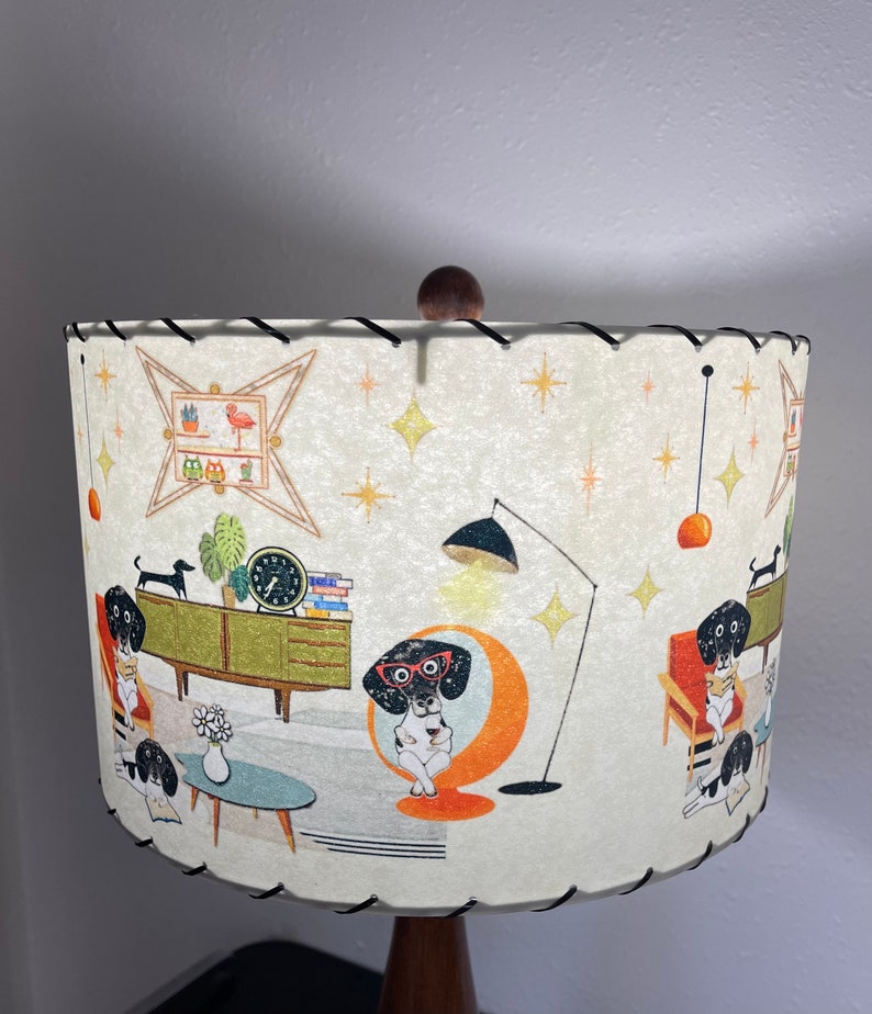 May include: A white lampshade with a black and white illustration of dogs in a mid-century modern living room. The lampshade has a black trim and a wooden finial.