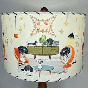May include: A white lampshade with a black trim features a mid-century modern design with three dachshunds, a potted plant, a clock, and books. The lampshade has a white background with gold stars and a black and white geometric pattern.