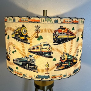 May include: A vintage lampshade with a cream background and a repeating pattern of colorful toy trains on a track. The trains are red, blue, green, yellow, and black. The lampshade has a scalloped edge and a black trim.