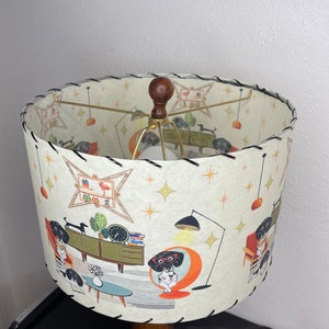 May include: A white lampshade with a black stitched edge features a mid-century modern design with illustrations of dogs in various poses. The lampshade has a gold metal frame and a brown wooden finial.