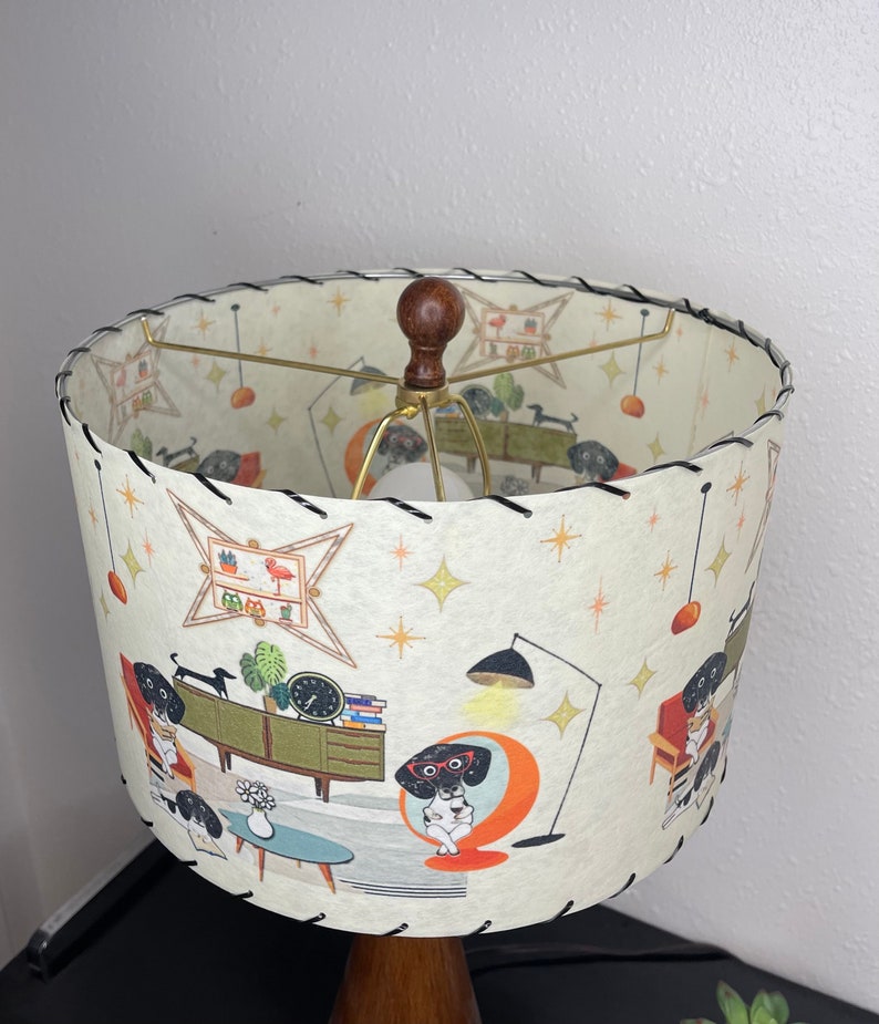 May include: A white lampshade with a black stitched edge features a mid-century modern design with illustrations of dogs in various poses. The lampshade has a gold metal frame and a brown wooden finial.