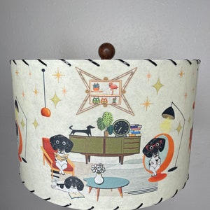 May include: A white lampshade with a black trim features a mid-century modern design with illustrations of dachshunds reading books in a living room setting. The lampshade has a brown wooden finial.