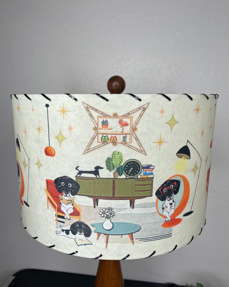 May include: A white lampshade with a black trim features a mid-century modern design with illustrations of dachshunds reading books in a living room setting. The lampshade has a brown wooden finial.