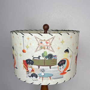 May include: A white lampshade with a black trim and a mid-century modern design featuring black and white dogs, a green plant, a clock, and a bookshelf with a pink flamingo. The lampshade is on a wooden base.